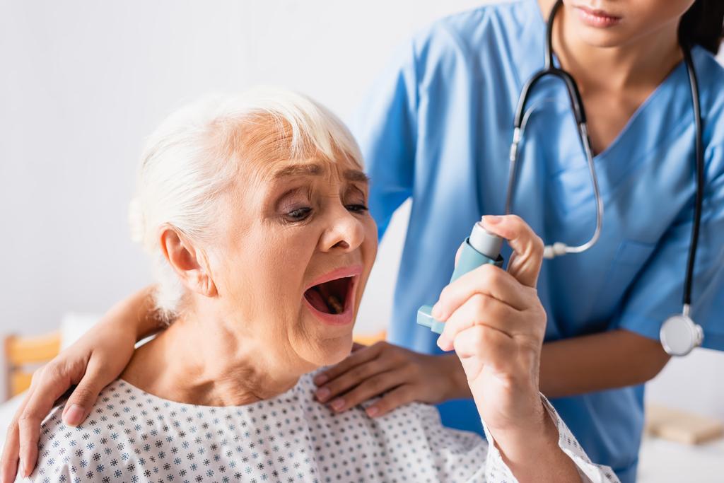 elderly woman using inhaler while suffering from asthma attack near nurse touching her shoulders on blurred background - Photo, Image