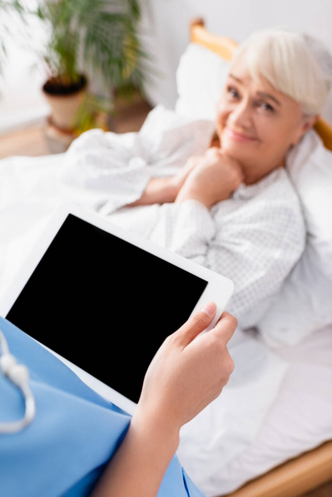 χαρούμενη ηλικιωμένη γυναίκα που κοιτάζει νοσοκόμα κρατώντας ψηφιακό δισκίο με κενή οθόνη, θολή φόντο - Φωτογραφία, εικόνα