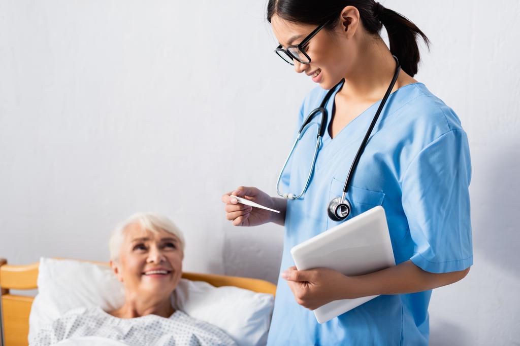 sonriente asiático enfermera con digital tableta mirando termómetro cerca feliz anciano mujer en borrosa fondo - Foto, Imagen
