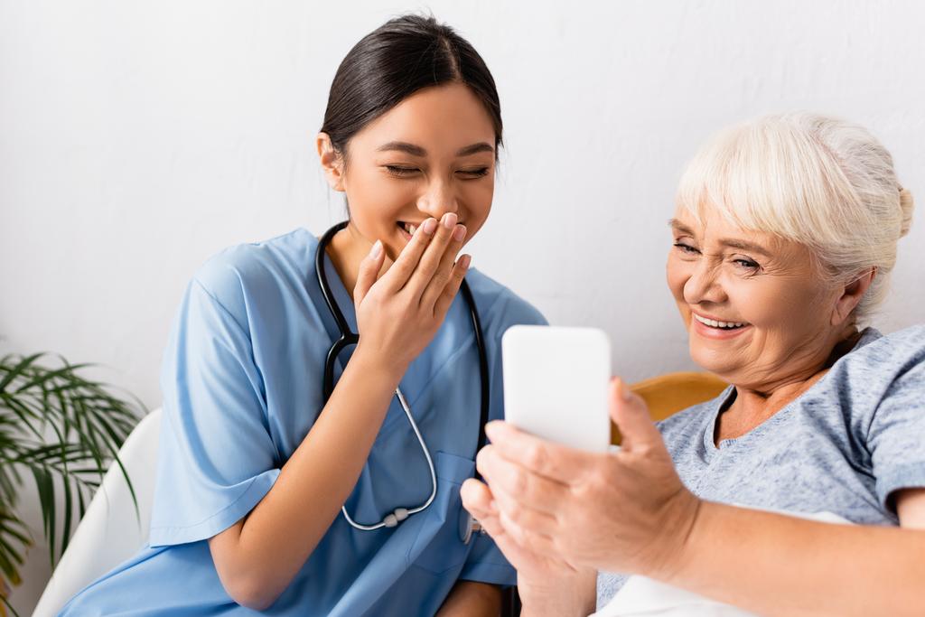 riendo asiático enfermera cubierta boca con mano cerca alegre senior mujer usando móvil teléfono, borrosa primer plano - Foto, Imagen
