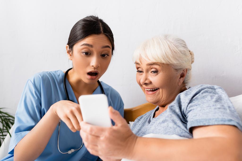 έκπληκτη Ασιάτισσα νοσοκόμα που δείχνει το κινητό της κοντά σε ενθουσιασμένες ηλικιωμένες γυναίκες, θολή πρώτο πλάνο - Φωτογραφία, εικόνα