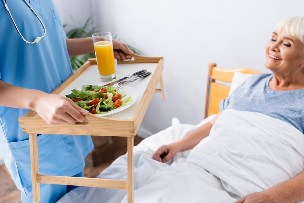 enfermera celebración bandeja con desayuno fresco cerca complacido mujer mayor, fondo borroso - Foto, imagen