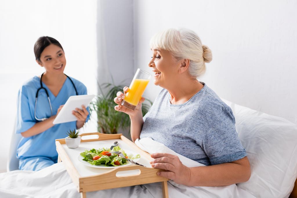 pleased aged woman having breakfast and drinking orange juice near smiling asian nurse on blurred background - Photo, Image