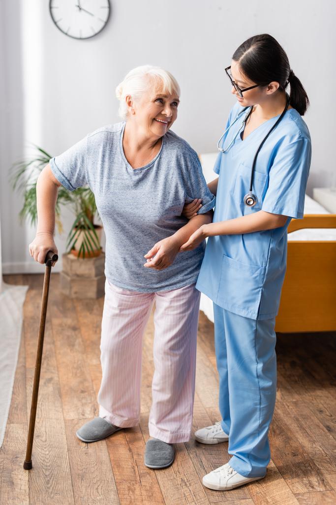 bejaarde vrouw met wandelstok kijken naar jong aziatisch verpleegster ondersteunen haar in ziekenhuis - Foto, afbeelding