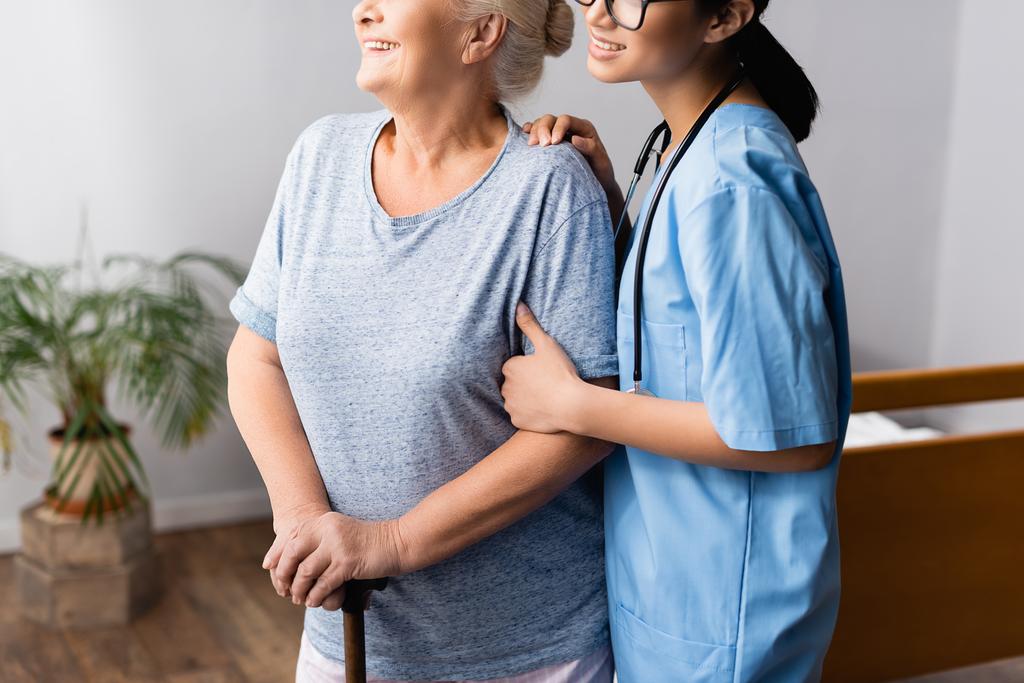 Hastanede bastonuyla yürüyen yaşlı bir kadını destekleyen gülümseyen hemşire görüntüsü - Fotoğraf, Görsel