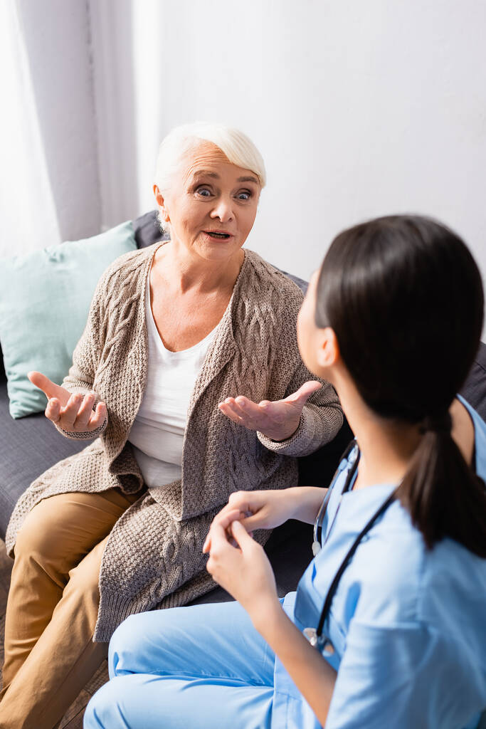 έκπληκτος ηλικιωμένη γυναίκα gesturing κατά τη διάρκεια της συνομιλίας με τη νοσοκόμα στο γηροκομείο, θολή πρώτο πλάνο - Φωτογραφία, εικόνα