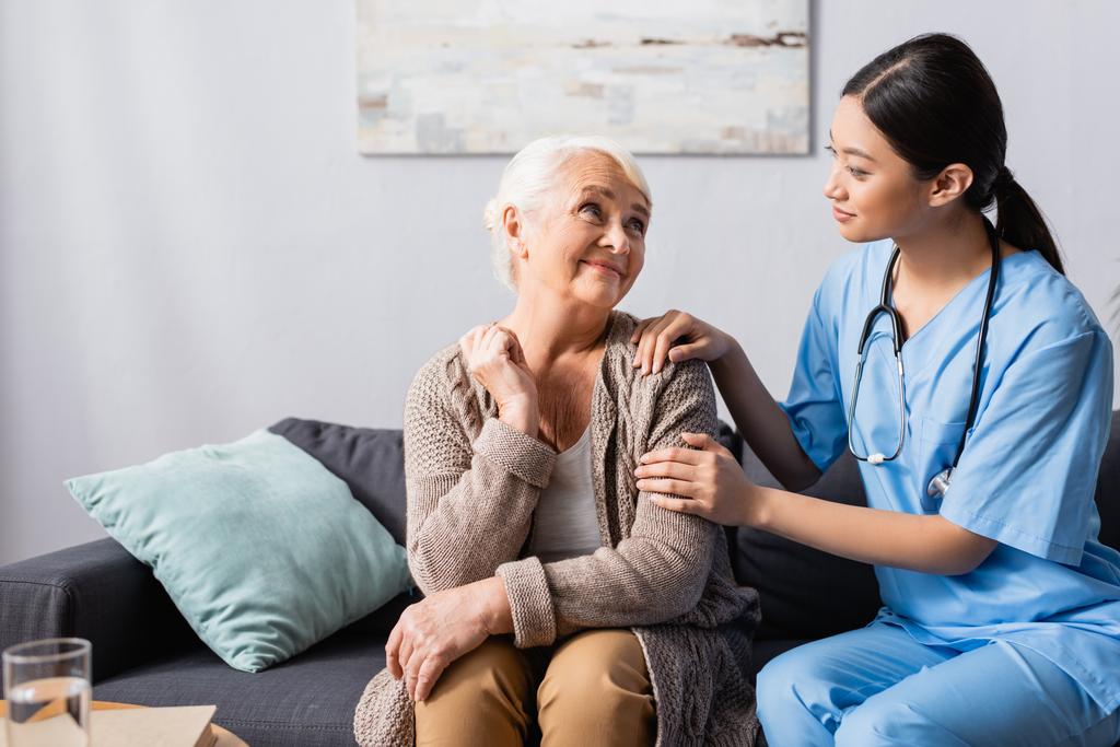 junge asiatische Krankenschwester berührt lächelnde Seniorin im Gespräch im Pflegeheim - Foto, Bild