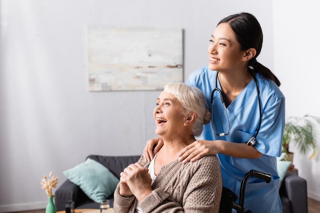 счастливая азиатская медсестра трогает плечи пожилой женщины-инвалида, смеющейся в инвалидном кресле - Фото, изображение