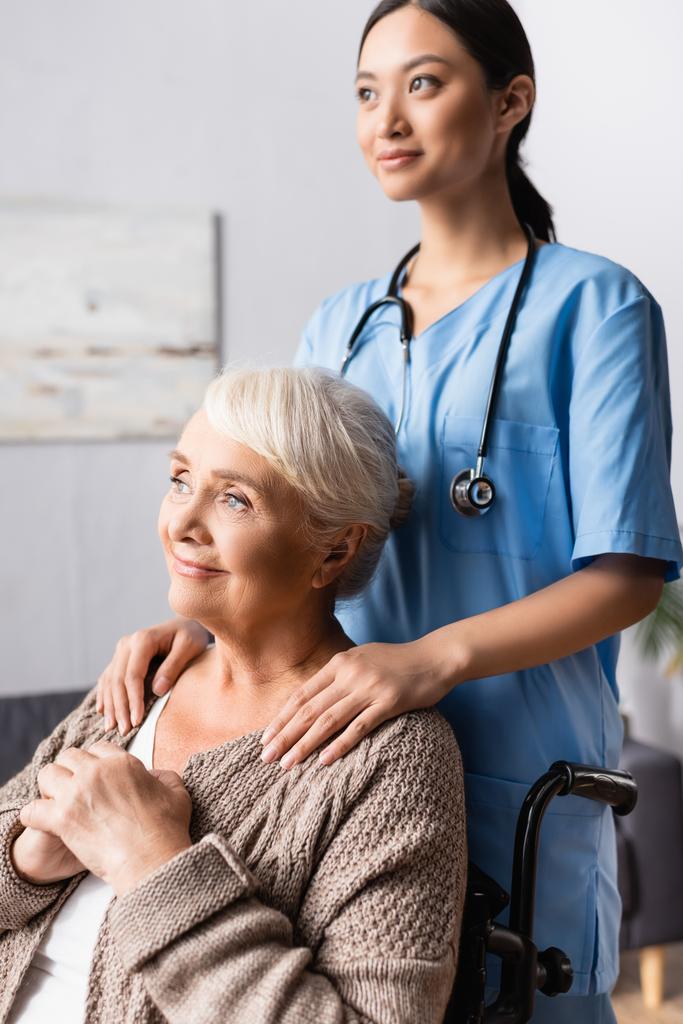 jovem asiático enfermeira tocando ombros de sorrir idosa mulher olhando afastado em cadeira de rodas - Foto, Imagem