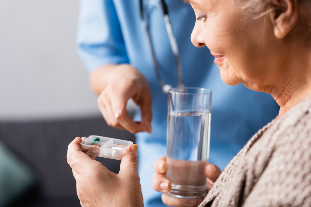 verpleegster wijzend met de vinger naar pillen in de hand van oudere vrouw met glas water, wazig voorgrond - Foto, afbeelding