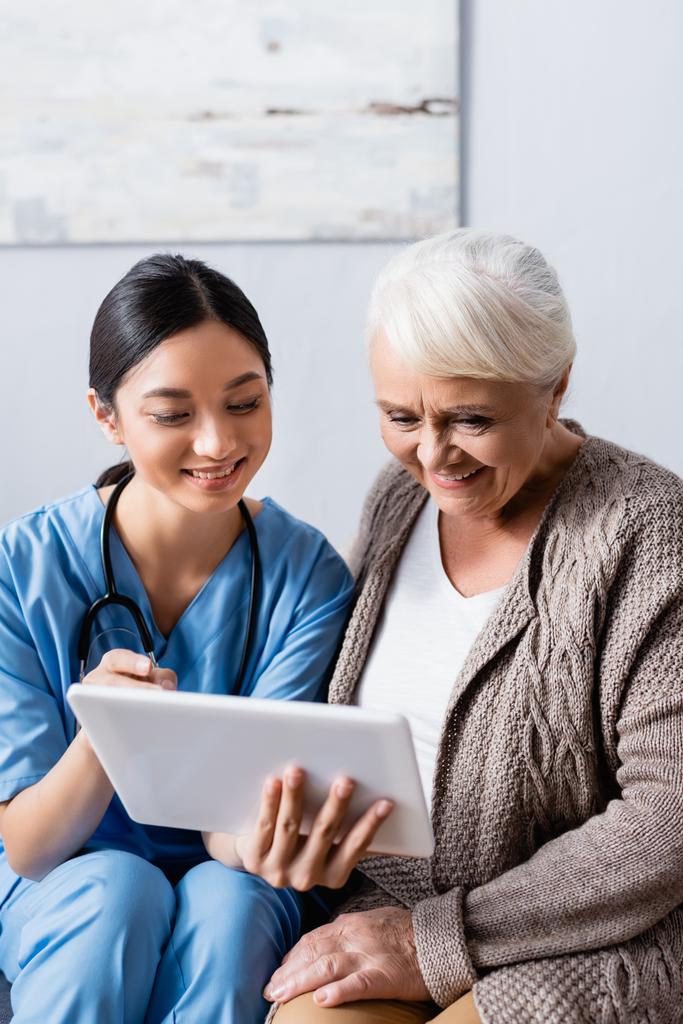wesoła azjatycka pielęgniarka wskazująca palcem na cyfrowy tablet w pobliżu uśmiechniętej starszej kobiety - Zdjęcie, obraz