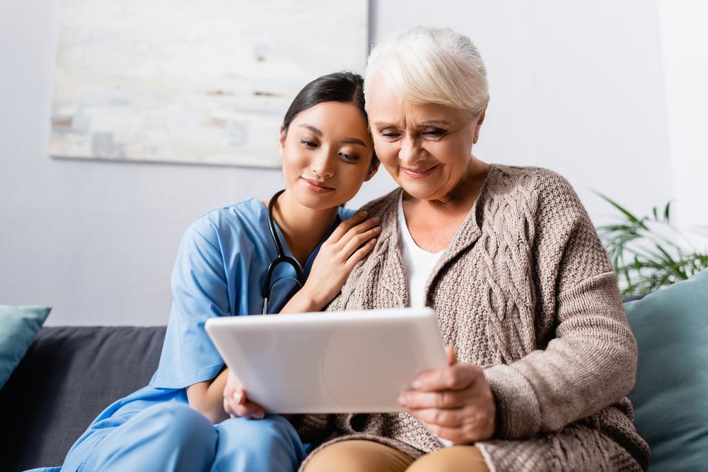 happy asian woman leaning on shoulder of aged smiling woman holding digital tablet - Photo, Image