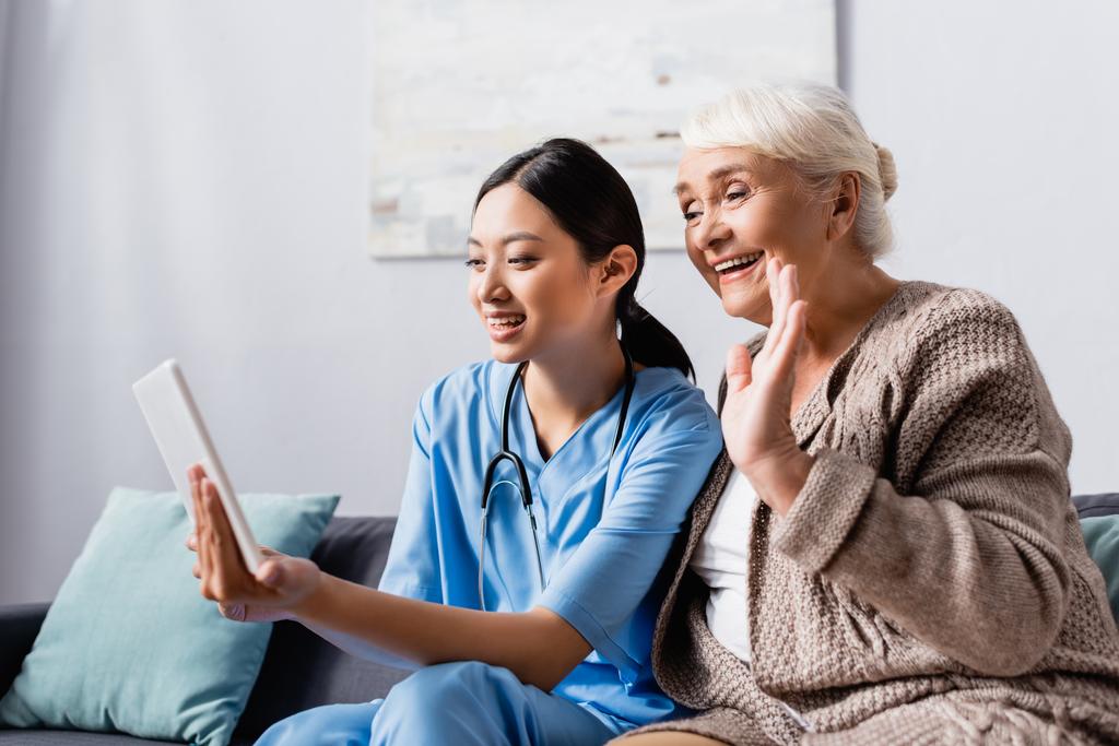 χαμογελαστή ασιατική νοσοκόμα κρατώντας ψηφιακή ταμπλέτα κοντά ευτυχισμένη ηλικιωμένη γυναίκα χαιρετώντας το χέρι κατά τη διάρκεια της ομιλίας βίντεο - Φωτογραφία, εικόνα