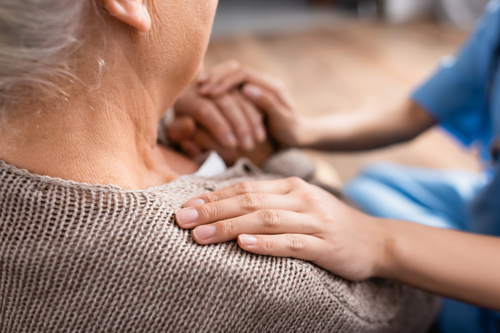 abgeschnittene Ansicht der Krankenschwester, die Hand und Schulter einer älteren Patientin im Pflegeheim berührt, verschwommener Hintergrund - Foto, Bild