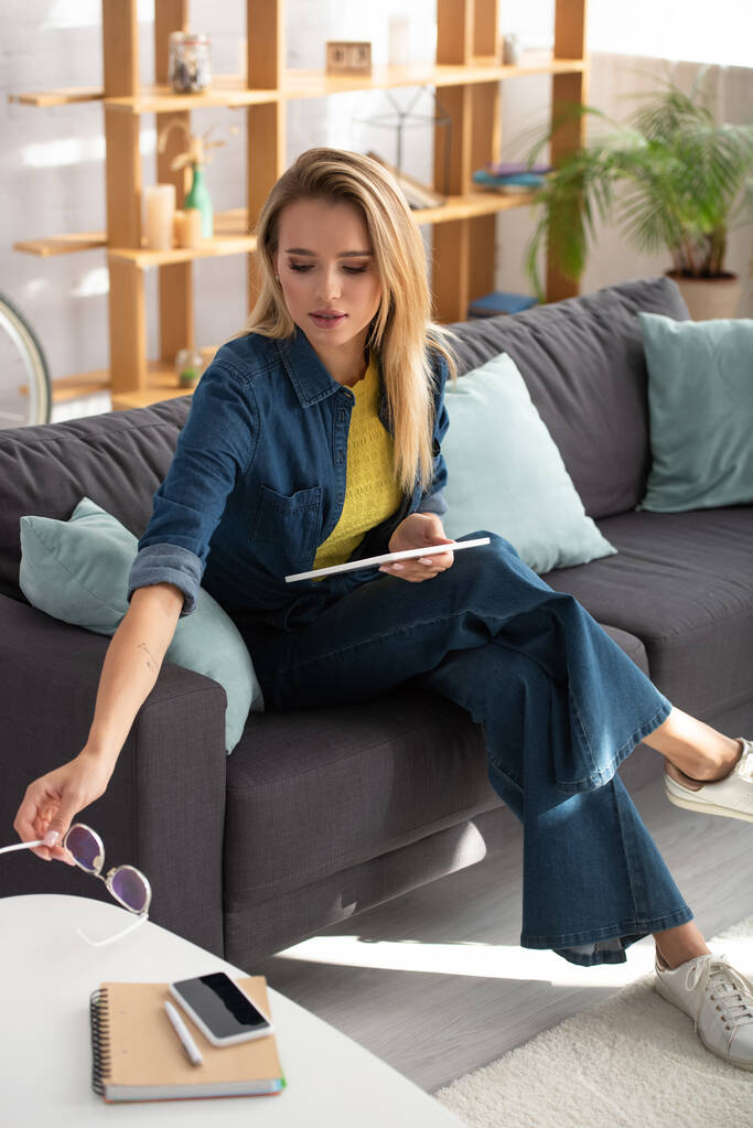 Young blonde woman with digital tablet putting eyeglasses on coffee table while sitting on couch on blurred background - Photo, Image