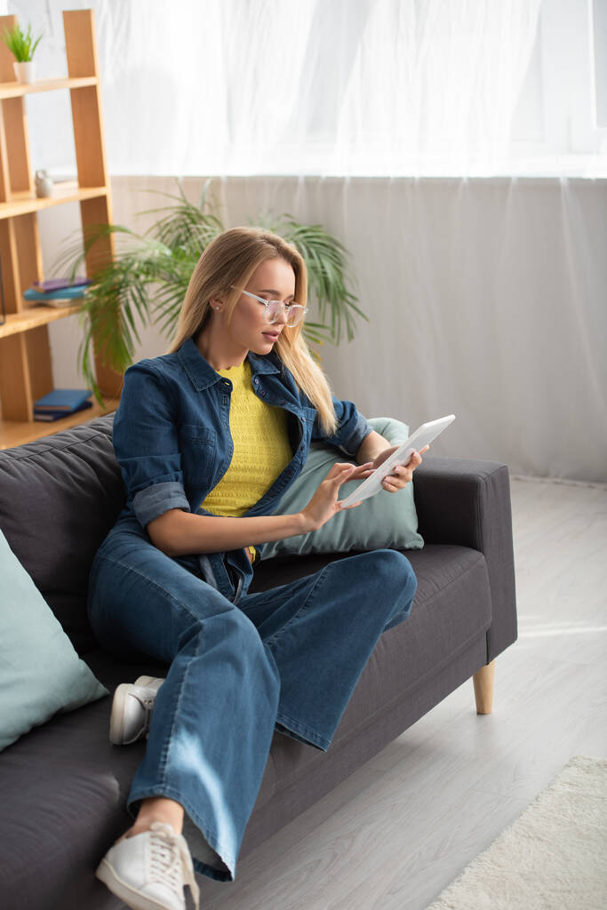 Comprimento total de jovem loira em óculos usando tablet digital enquanto sentado no sofá em fundo borrado - Foto, Imagem