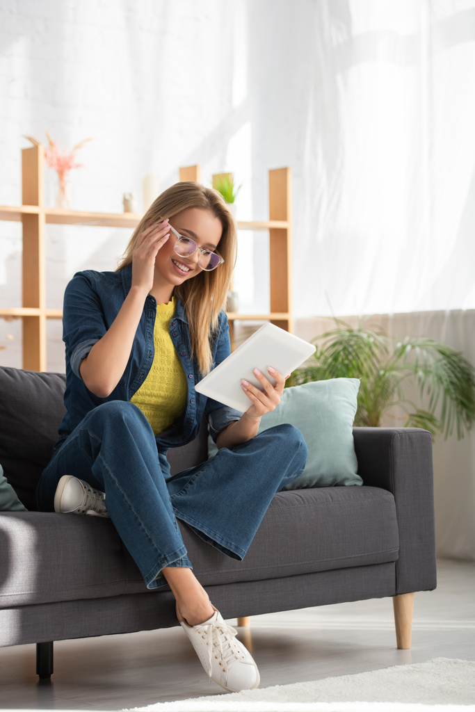 Volle Länge einer lächelnden jungen Frau mit Brille, die auf dem Sofa vor verschwommenem Hintergrund auf ein digitales Tablet blickt - Foto, Bild