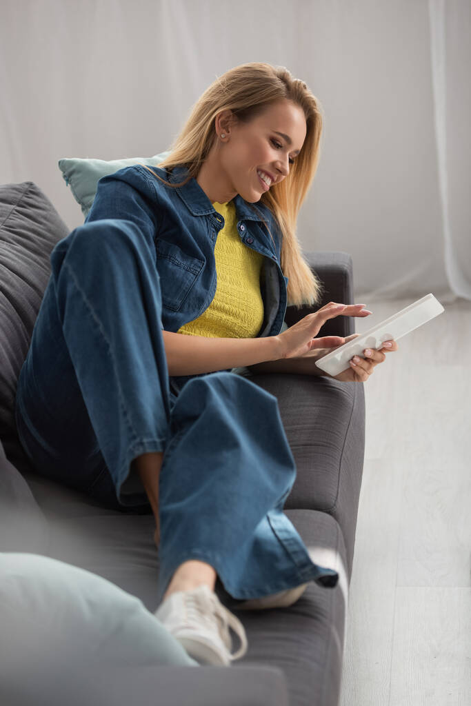Uśmiechnięta blondynka korzystająca z tabletu cyfrowego leżąca na kanapie w domu na niewyraźnym pierwszym planie - Zdjęcie, obraz