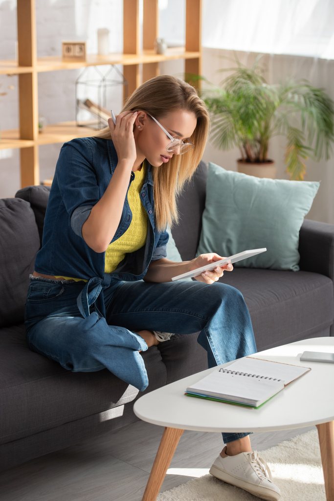 Longitud completa de mujer rubia joven mirando tableta digital mientras está sentado en el sofá cerca de la mesa de café sobre fondo borroso - Foto, Imagen