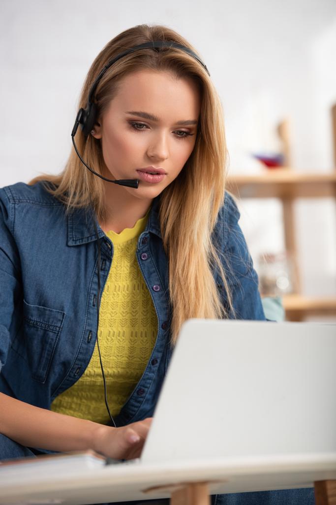 Young blonde woman in headset looking at laptop at home on blurred background - Photo, Image