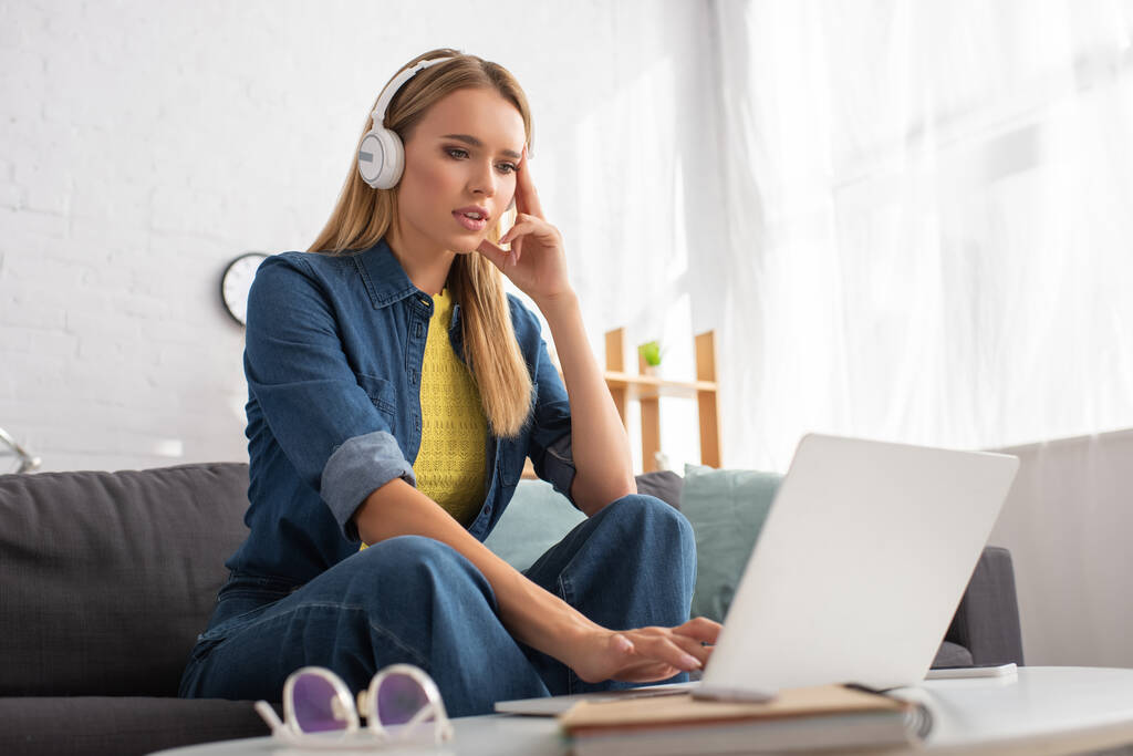 Bedachtzame jonge vrouw in hoofdtelefoon met behulp van laptop terwijl zitten op de bank thuis op wazig voorgrond - Foto, afbeelding
