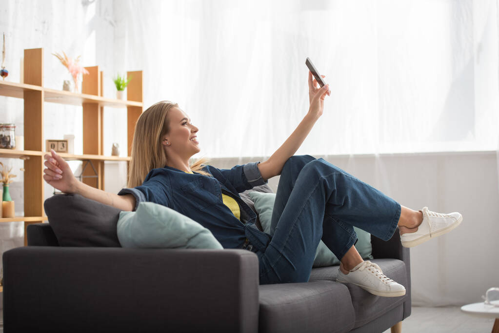 Piena lunghezza di donna bionda sorridente con smartphone scattare selfie mentre seduto sul divano su sfondo sfocato - Foto, immagini