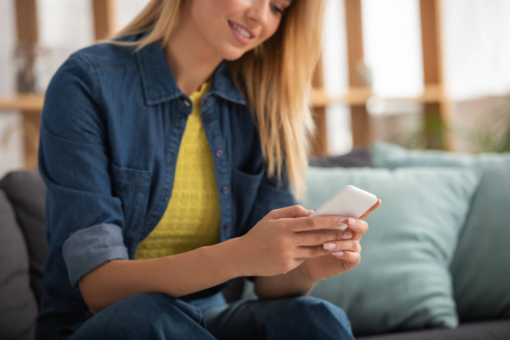 Обрізаний вид на молоду блондинку, що пише на смартфоні, сидячи на дивані на розмитому фоні
 - Фото, зображення