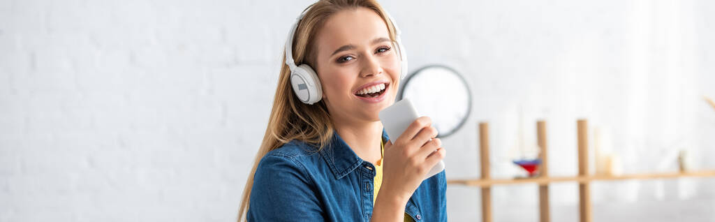 背景がぼやけている自宅でスマートフォンを保持しながら、ヘッドフォンの歌で笑顔の若い女性,バナー - 写真・画像