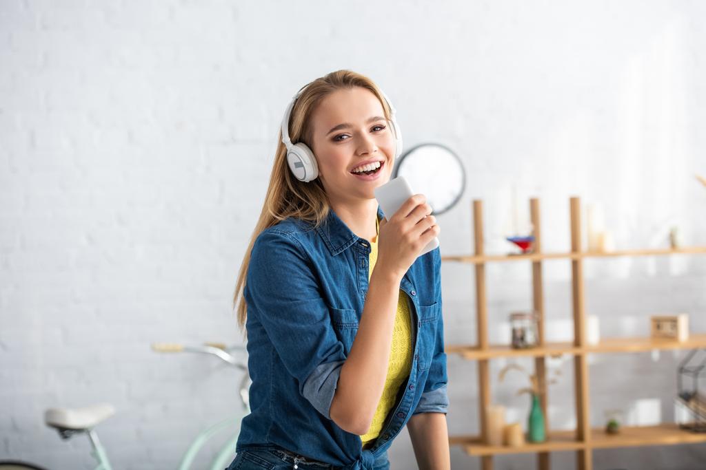 Mujer rubia alegre con teléfono inteligente mirando a la cámara mientras canta en casa sobre fondo borroso - Foto, Imagen