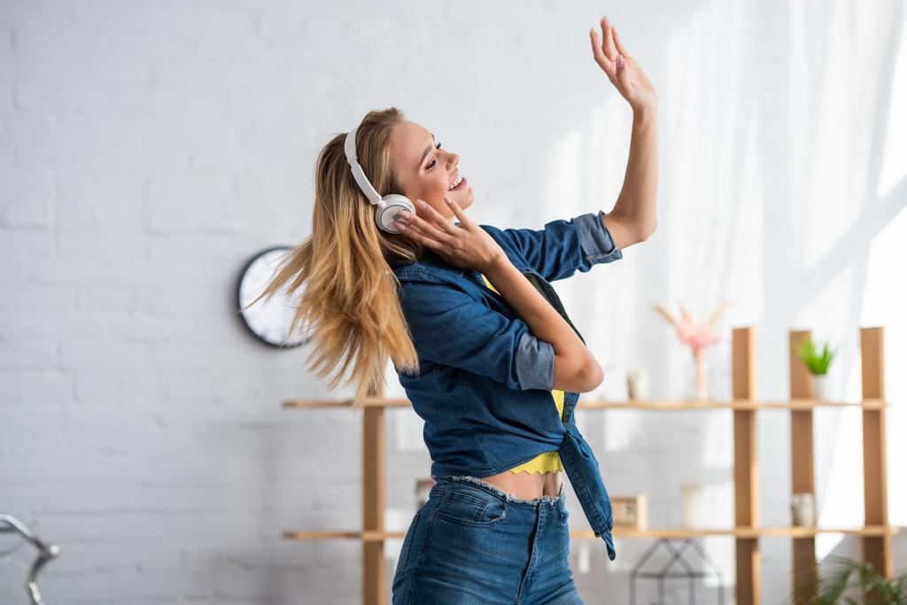 Wesoła młoda kobieta w słuchawkach tańcząca w domu na rozmytym tle - Zdjęcie, obraz