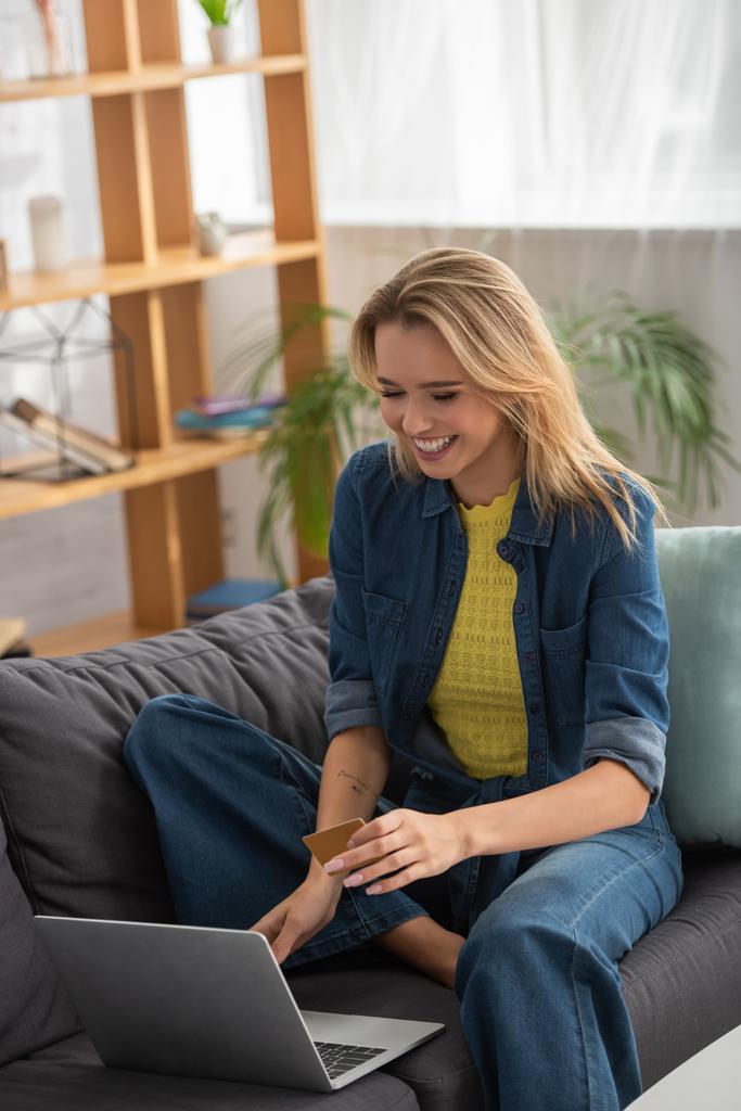 Happy blonde woman with credit card laughing while sitting on couch near laptop on blurred background - Photo, Image