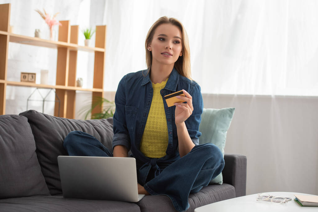 Nuori blondi nainen luottokortilla katselee pois istuessaan läppärin lähellä sohvalla hämärtyneellä taustalla - Valokuva, kuva