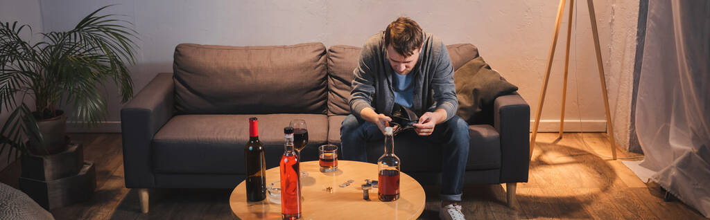 μεθυσμένος άνδρας κρατώντας άδειο πορτοφόλι κοντά στο τραπέζι με μπουκάλια αλκοολούχων ποτών στο σπίτι, πανό - Φωτογραφία, εικόνα