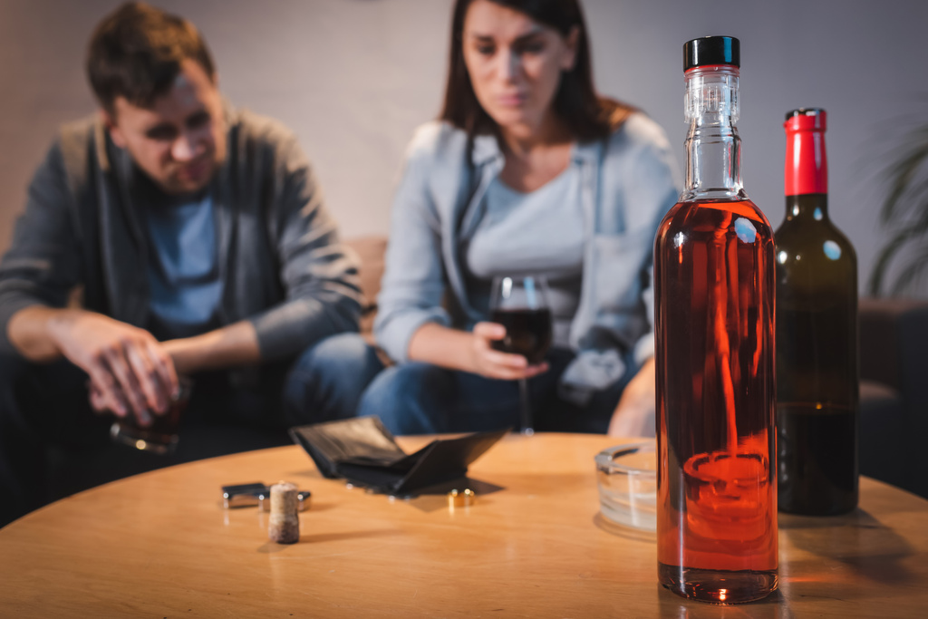 bottles of alcohol and empty wallet on table near drunk couple on blurred background - Photo, Image