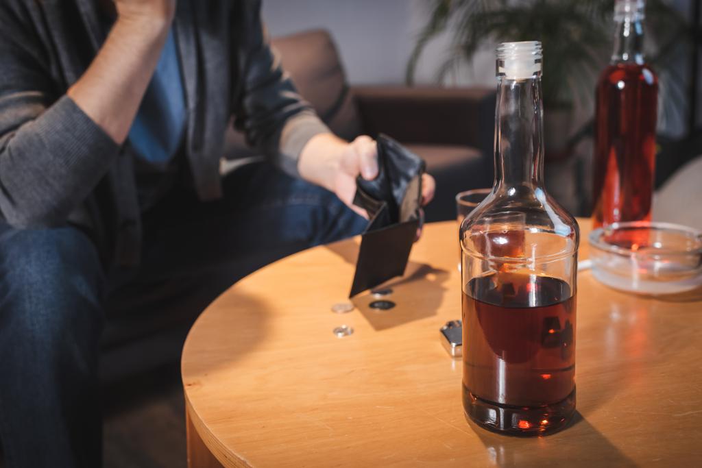 vista cortada de homem viciado segurando carteira vazia perto de garrafas com álcool, fundo borrado - Foto, Imagem