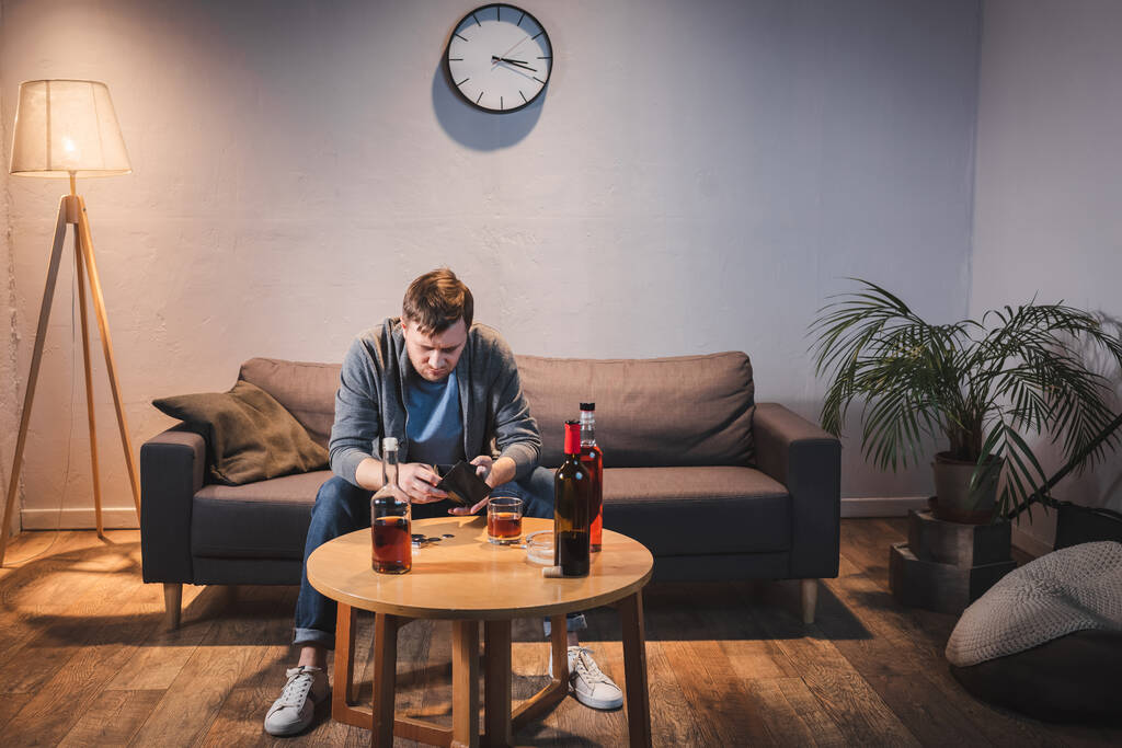 addicted man holding empty wallet near bottles of alcohol drinks on table - Photo, Image