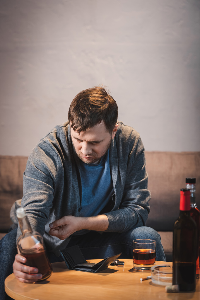 frustrated, addicted man holding bottle of whiskey near empty wallet on table - Photo, Image