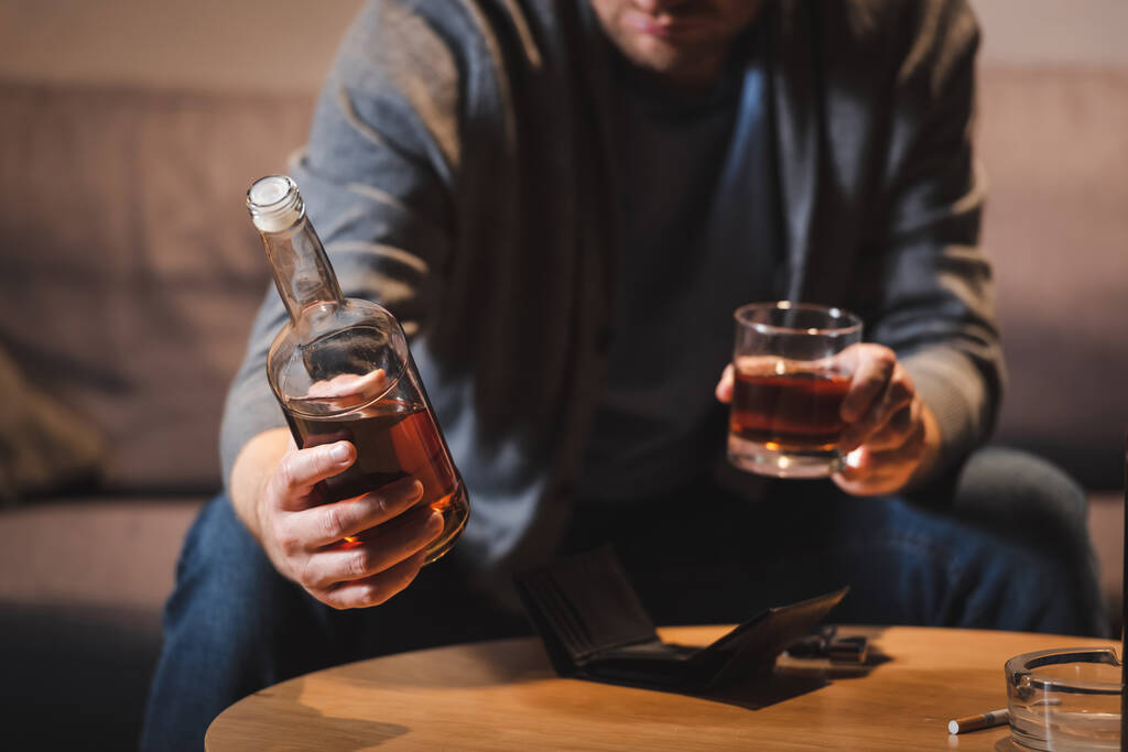 vista recortada del hombre alcohólico sosteniendo botella y vaso de whisky en casa, fondo borroso - Foto, imagen