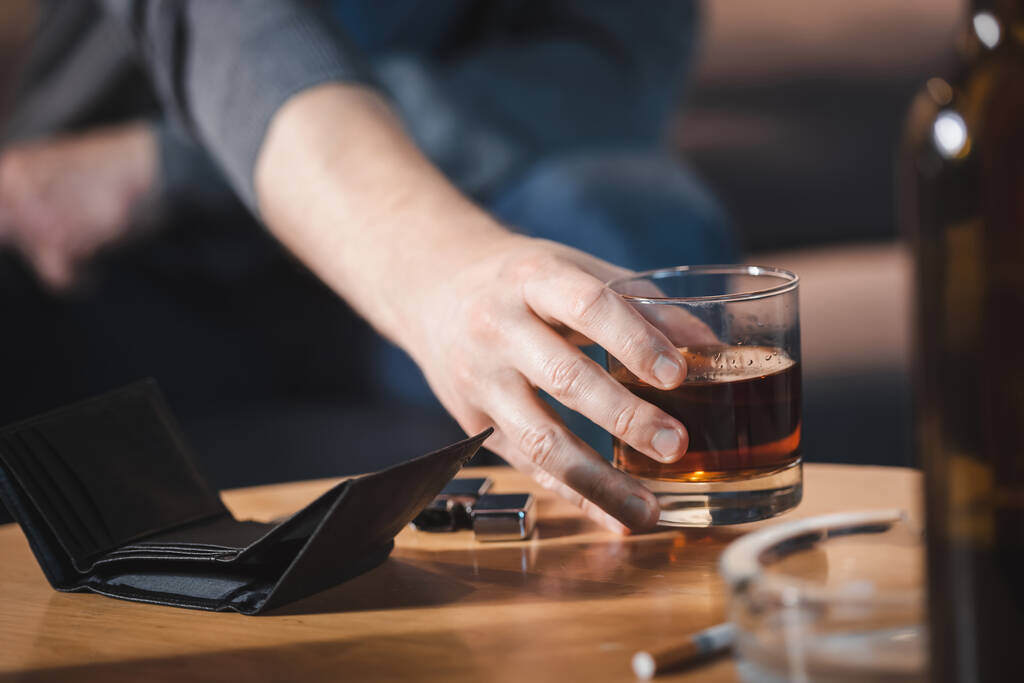 enfoque selectivo del hombre adicto tomando vaso de whisky cerca de la cartera vacía en primer plano borrosa - Foto, Imagen
