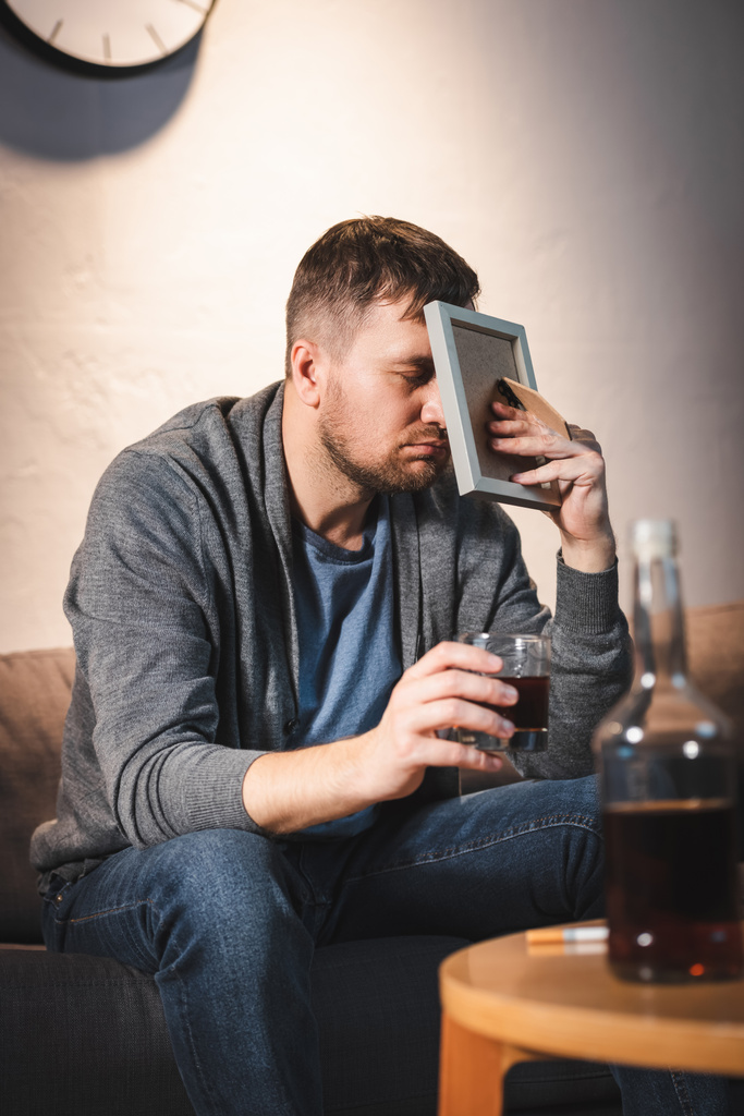 Depressiver Mann verdeckt Gesicht mit Fotorahmen, während er ein Glas Whiskey in der Hand hält - Foto, Bild