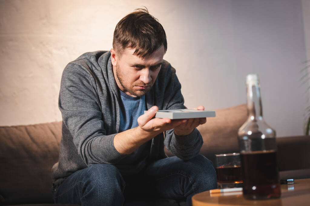 depresivní muž sedí u stolu s alkoholickými nápoji a drží fotorámeček - Fotografie, Obrázek