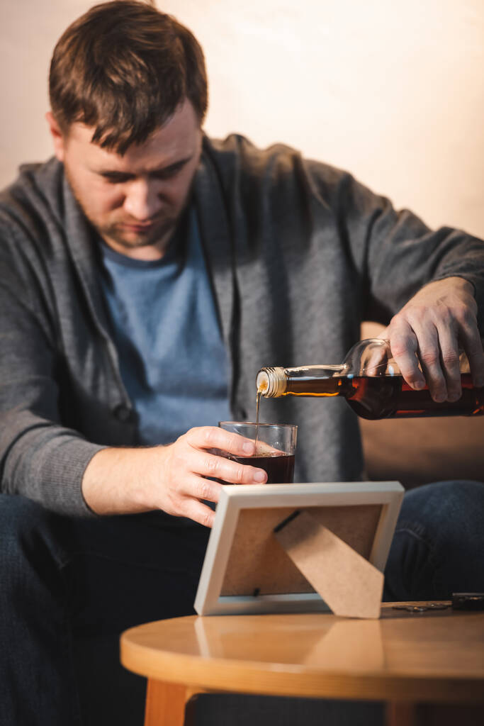 addicted man pouring whiskey into glass near photo frame, blurred background - Photo, Image