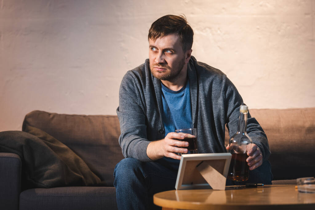 depressed man holding glass and bottle of whiskey near photo frame on table - Photo, Image