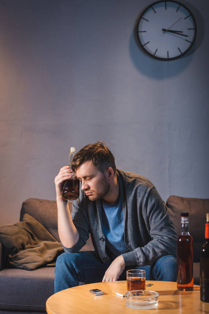 Betrunkener hält Flasche Alkohol in der Hand, während er allein zu Hause sitzt - Foto, Bild