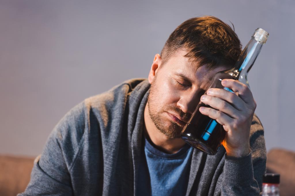 Alkoholsüchtiger Mann sitzt mit Flasche Whiskey am Kopf mit geschlossenen Augen - Foto, Bild