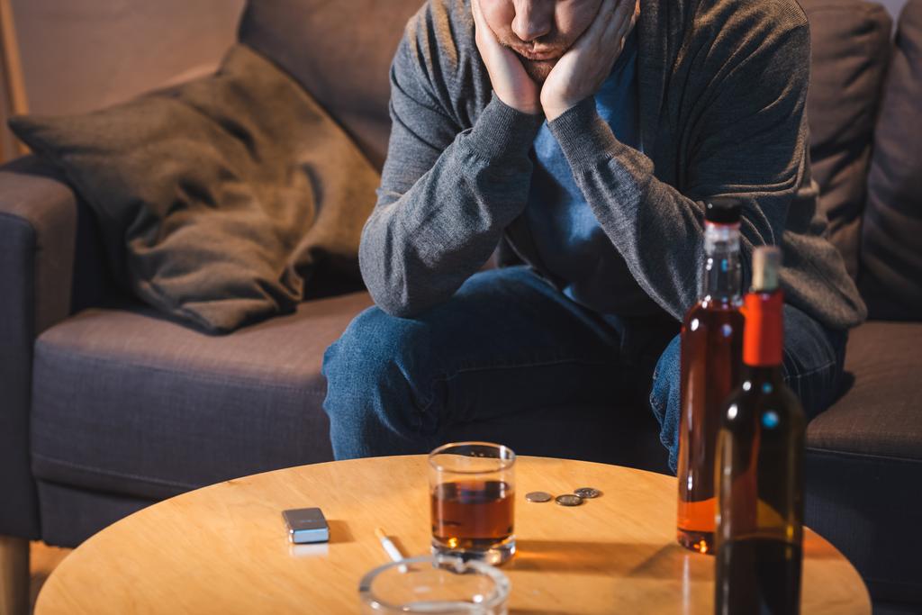 homem bêbado sentado perto da mesa com vidro e garrafas de álcool em casa - Foto, Imagem