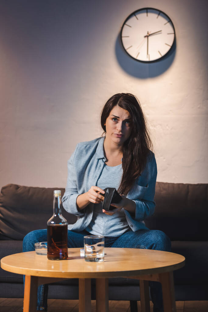 Betrunkene Frau blickt in Kamera und hält leere Geldbörse neben Whiskey-Flasche auf Tisch - Foto, Bild