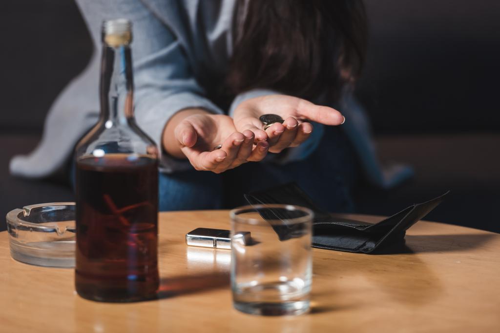 częściowy widok kobiety alkoholiczki trzymającej monety w pobliżu whisky i pusty portfel na stole, niewyraźne tło - Zdjęcie, obraz