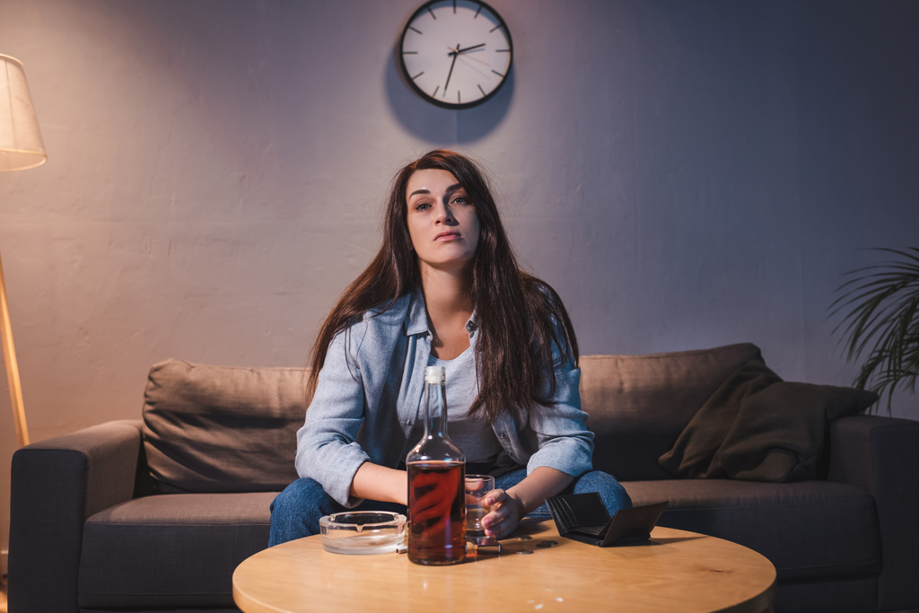 mujer adicta al alcohol sentada cerca de la botella de whisky y la cartera vacía en casa   - Foto, imagen