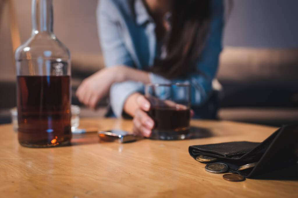 oříznutý pohled na ženu závislou na alkoholu, jak si bere sklenici whisky s mincemi poblíž peněženky, rozmazané pozadí - Fotografie, Obrázek